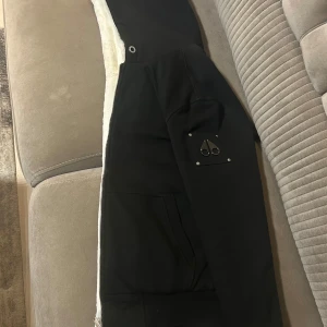 Svart pufferjacka från Moose Knuckles - Svart pufferjacka från Moose Knuckles med vit fuskpäls på insidan av huvan och quiltat innerfoder. Jackan har dragkedja framtill, två fickor och logotyp på ärmen. Perfekt för kalla dagar och har en modern, stilren look.