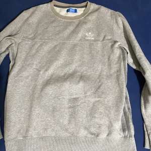 Grå sweatshirt från Adidas Originals - Snygg grå sweatshirt från Adidas Originals med diskret logga på bröstet. Tröjan har rund hals med ribbad kant och långa ärmar. Mjuk och skön bomullsblandning, perfekt för chill eller träning. Klassisk och enkel design som funkar till allt.