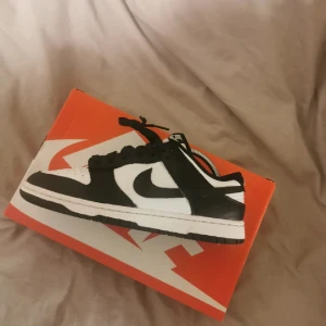 Nike Dunk Low svart/vit sneaker - Säljer en klassisk Nike Dunk Low i svart och vitt med tydlig Swoosh-logga på sidan. Skon har snörning, platt sula och rund tå. Perfekt för dig som gillar streetstyle och vill ha en ikonisk sneaker i garderoben.