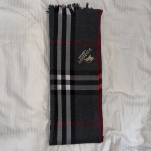 Rutig halsduk från Burberry London - Snygg halsduk från Burberry London med klassiskt rutigt mönster i mörkgrått, svart, vitt och röda detaljer. Broderad logga med riddare på ena sidan. Mjuk och stilren, perfekt för att lyfta din outfit under kyliga dagar.