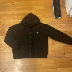 Svart hoodie från Polo Ralph Lauren - Svart hoodie från Polo Ralph Lauren i custom fit med dragkedja framtill och klassisk huva. Tröjan har lång ärm och den ikoniska vita Polo-loggan broderad på bröstet. Perfekt för chill dagar och enkel att matcha med jeans eller joggers.