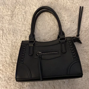 Svart handväska med dragkedja - Snygg svart handväska i skinnimitation med både handtag och avtagbar axelrem. Väskan har ett stilrent, rektangulärt format och dekorativa sömmar på sidorna. Framtill finns en liten ficka med dragkedja och långa tofsar som detalj. Perfekt storlek för det viktigaste.