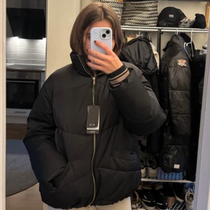 Svart pufferjacka från Armani Exchange - Svart pufferjacka från Armani Exchange med hög krage, guldfärgad dragkedja och ribbade muddar med gråa ränder. Jackan har två fickor framtill och quiltad design. Perfekt för kalla dagar och riktigt snygg till streetwear.