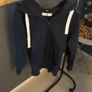 Mörkblå hoodie från Russermerch  - Mörkblå hoodie från Russermerch i storlek S med vit snörning och stor huva. Tröjan har praktisk dragkedja framtill och en stor ficka på magen. Perfekt för chill dagar och enkel att matcha med jeans eller joggers.