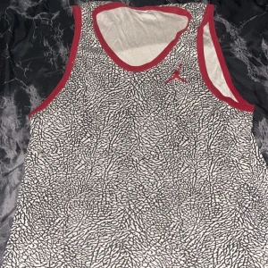 Jordan linnetopp med grafiskt mönster - Säljer en snygg linnetopp från Jordan med ett grafiskt svartvitt mönster och röda kanter. Toppen har ärmlös design och ett litet rött Jumpman-logo på bröstet. Perfekt för dig som gillar sportig streetstyle och vill sticka ut.