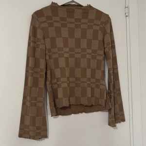 Brun rutig långärmad topp - Säljer en brun långärmad topp med rutigt mönster i två nyanser av brunt. Toppen har en lätt croppad passform med vågig kant vid ärmslut och nederkant. Perfekt för dig som gillar retrovibbar och unika detaljer.