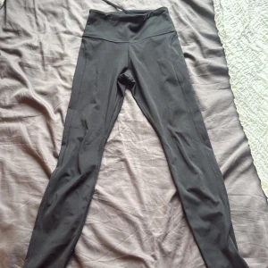 Svarta Nike Dri-Fit leggings - Svarta leggings från Nike med Dri-Fit-teknologi, perfekt för träning. Hög midja och tight passform, med meshpaneler på baksidan av benen för extra ventilation. Materialet är syntetiskt och stretchigt för maximal komfort under rörelse.