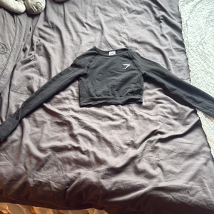 Svart croppad långärmad topp Gymshark - Säljer en svart croppad långärmad topp från Gymshark i storlek XS. Toppen har ett diskret Gymshark-logotryck på bröstet och är tillverkad i ett stretchigt syntetmaterial med ribbad struktur. Perfekt för dig som gillar sportig och stilren look.