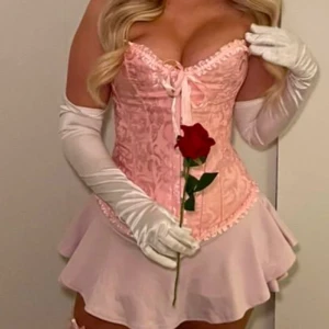 Rosa korsett med snörning och rosett - Säljer en superfin rosa korsett med glansigt mönster, snörning i ryggen och söt rosett framtill. Korsetten har volangkant upptill och nedtill, samt metallknappar framtill. Materialet är glansigt och känns lyxigt, perfekt för att skapa en statement-look.