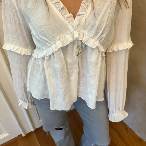 Vit blus med volang och v-ringning - Supersöt vit blus med v-ringning och volangdetaljer längs halsringning, ärmslut och över bysten. Långärmad och lätt transparent med struktur i tyget. Perfekt att styla med jeans för en chill men ändå romantisk look.