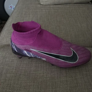 Nike Phantom GT2 rosa fotbollsskor - Pris kan diskuteras. Nike Phantom GT2 fotbollsskor i en snygg rosa färg med svart och vitt blixtmönster på hälen. Skorna har en strumpliknande passform utan synlig snörning, stor svart Swoosh på sidan och textilöverdel för bra bollkontroll. Perfekta för dig som vill sticka ut på planen. Dem är helt nya används 1 gång pirs kan diskuteras 