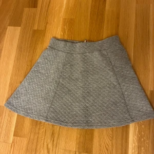 Grå quiltad kortkjol från Club XPRS - Snygg grå kortkjol från Club XPRS med quiltad struktur och markerad midja. Kjolen har en synlig dragkedja bak och är tillverkad i mjuk bomull och polyesterblandning. Perfekt för dig som gillar stilrena och bekväma plagg med lite edge.