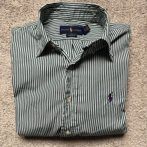 Randig skjorta från Ralph Lauren - Snygg klassisk skjorta från Ralph Lauren i grön och vit randigt mönster. Classic fit med lång ärm, krage och knappar framtill. Broderad lila logga på bröstet ger en extra touch. Tillverkad i mjuk bomull som känns skön mot huden. Är grön.