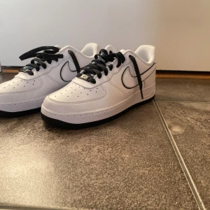 *Oanvända* Nike Air Force 1 sneakers - Nike Air Force 1 sneakers i vitt skinn med svarta detaljer och snörning. Klassisk rund tå och platt sula, grön Nike-logga på hälen och svart swoosh med vit kant. Perfekt för dig som gillar stilrena och tidlösa sneakers med streetkänsla. Köpare betalar frakt.