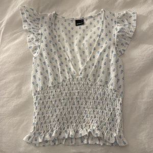 Vit blommig blus från Gina Tricot - Supersöt blus från Gina Tricot i vit med ljusblått blommönster. Toppen har v-ringad hals, korta volangärmar och smockad midja med volangkant nedtill. Materialet är mjukt och lätt, perfekt för varma dagar. Säljer då den inte har kommit till användning någon gång!