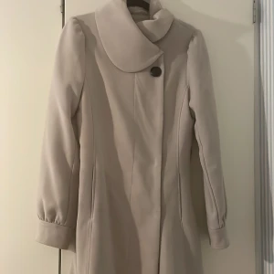 Beige kappa från H&M med stor krage - Snygg beige kappa från H&M i storlek 36. Jackan har en stor, rundad krage och stängs med dolda tryckknappar. Dekorativ synlig knapp framtill. Långa ärmar med lätt puff vid axlarna och markerad midja baktill. Hällor finns om du vill addera ett skärp. Perfekt för dig som gillar stilrena och klassiska plagg.