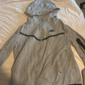 Grå hoodiejacka från Nike XL - Grå hoodiejacka från Nike i storlek XL. Jackan har hel dragkedja, huva och svarta detaljer som en rand över bröstet och en ficka på ärmen. Tillverkad i mjuk bomullsblandning, perfekt för chill dagar eller när du är på språng.