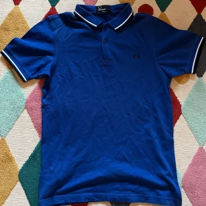 Blå pikétröja Fred Perry slim fit - Snygg blå pikétröja från Fred Perry i slim fit-modell. Tröjan har klassisk krage med vita ränder och korta ärmar med matchande vit kant. Broderad Fred Perry-logga på bröstet. Tillverkad i mjuk bomull som är skön mot huden. Använd 1-3 gånger!