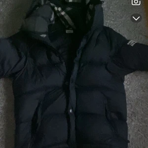 Svart pufferjacka från Burberry M - Säljer en svart pufferjacka från Burberry i storlek M. Jackan har klassisk Burberry-rutig huva, dragkedja och knappar framtill samt vadderad design som håller dig varm. Perfekt för kalla dagar och har en snygg, stilren look.