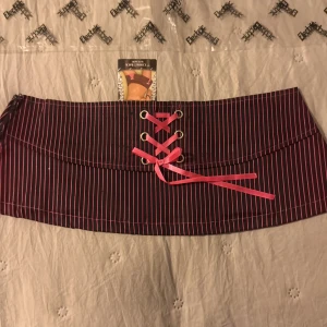 Randig minikjol från Electric Lingerie - Svart minikjol med tunna rosa ränder från Electric Lingerie. Kjolen har snörning framtill med rosa band och små rosetter som detaljer. Tillverkad i mjuk bomull med lite stretch för en bekväm passform. Perfekt för dig som gillar edgy och lekfull stil.