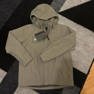 Beige regnjacka från Didriksons - Helt ny med prislap/ Snygg beige regnjacka från Didriksons med huva och diskret logga på bröstet. Jackan är 100% vattentät och har en stilren design med dragkedja framtill. Perfekt för blöta dagar och enkel att matcha med din vardagsstil.