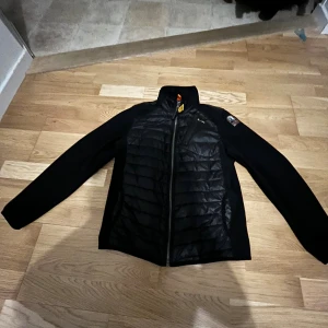 Svart quiltad jacka Parajumpers Y-M - Svart jacka från Parajumpers med quiltad front och släta ärmar. Jackan har dragkedja, hög krage och coolt PJS-märke på ärmen. Materialet är polyester och syntet, perfekt för kyliga dagar. Orange och gul detalj vid kragen ger extra stilpoäng.