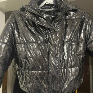 Svart pufferjacka från Boohoo - Säljer en svart, glansig pufferjacka från Boohoo i storlek 36. Jackan har quiltad design, huva, dragkedja och tryckknappar framtill. Perfekt för dig som vill ha en cool och varm jacka med streetkänsla. Materialet är syntet och jackan har praktiska fickor. Cropped puffer jacket.