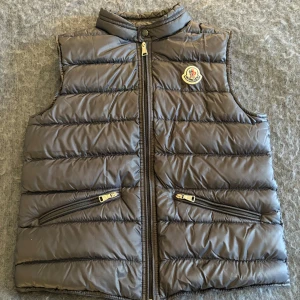 Svart dunväst från Moncler - Storlek 12/152 Cm. Snygg svart dunväst från Moncler med dragkedja och två fickor framtill. Västen har en hög krage och passar perfekt till lite kyligare sommar/vår dagar. Jättebra kvalitet, inte några skador eller något liknande. 