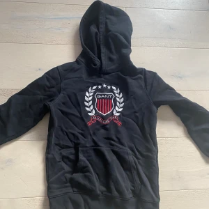 Svart hoodie från GANT med brodyr - Svart hoodie från GANT med stor broderad logga och sköld på bröstet i vitt och rött. Tröjan har huva, känguruficka och ribbade muddar. Perfekt för dig som gillar klassisk college-stil och vill ha något bekvämt och snyggt.