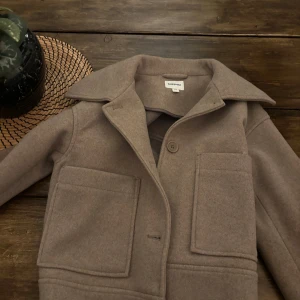 Beige kappa från Kappahl XS - Stilren beige overshirt från Kappahl i storlek XS. Jackan har stora framfickor, klassisk krage och knäppning med tre knappar framtill. Tillverkad i ett mjukt ullblandat material som passar perfekt till lager-på-lager. Snygga detaljer vid ärmslut med knapp.aldrig använd och köpte för 900kr!