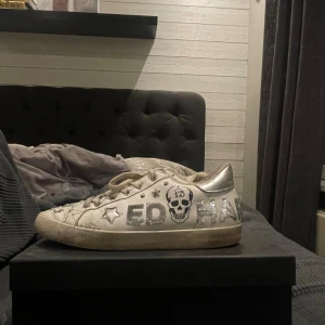 Ed Hardy vita sneakers - Unika vita och silverfärgade sneakers från Ed Hardy med coola detaljer som dödskalle, stjärnor och nitar. Skorna har snörning, låga skaft och röd sula. Perfekta för dig som vill sticka ut med en retro vibe och edgy look.