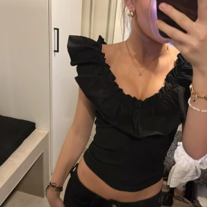 Svart volangtopp med djup ringning - Säljer en svart topp med bred volang runt urringningen och axlarna. Toppen är croppad och har en djup v-ringning både fram och bak. Perfekt för dig som gillar en feminin och trendig stil. Materialet känns som bomull eller polyester.