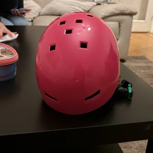 Rosa hjälm med tvättbjörnsdekal 52/56 - Cool rosa hjälm med fyrkantiga ventilationshål och justerbart spänne. På sidan finns en söt tvättbjörnsdekal med solglasögon och godis. Storlek 52/56, perfekt för cykel eller skateboard. Hjälmen har en modern, rundad form och är lätt att justera för bästa passform.