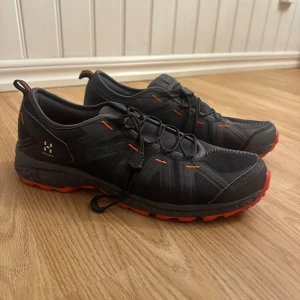 Svarta Haglöfs sneakers med orange detaljer - Snygga svarta sneakers från Haglöfs med orange detaljer och sula. Skorna har mesh- och syntetmaterial, snabbsnörning och sportig design. Perfekta för dig som gillar outdoor och vill ha en cool look med extra färg på fötterna. Aldrig använda endast provade inomhus 