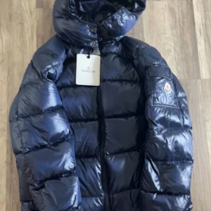 Mörkblå Moncler pufferjacka med huva - Säljer en snygg mörkblå pufferjacka från Moncler med glansig finish och brunt foder. Jackan har huva, dragkedja, fickor med dragkedja och Moncler-logga på ärmen. Perfekt för dig som vill ha en varm och stilren jacka med exklusiv känsla. Det följer inte med några skador från jackan. Priset kan sänkas vid snabb affär 