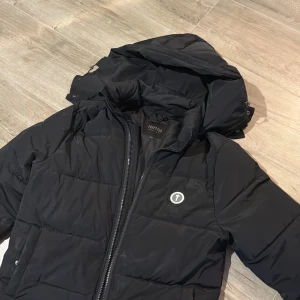 Svart pufferjacka från Trapstar - Säljer en svart pufferjacka från Trapstar med avtagbar huva och dragkedja framtill. Jackan har Trapstar-logga på bröstet och stor broderad text på ryggen. Perfekt för kalla dagar och riktigt street i stilen.