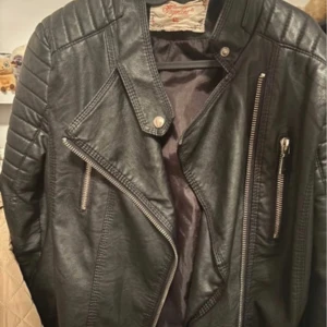 Svart skinnjacka med dragkedjor - Snygg svart skinnjacka från Freaky Nation med coola detaljer som snedställda dragkedjor och quiltade axelpartier. Jackan har en klassisk bikerstil med bred krage och metallknappar. Perfekt för dig som vill ha en edgy look.