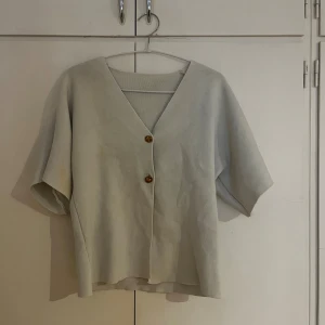Vit kofta med v-ringning och knappar - Snygg vit kofta med v-ringad hals och två bruna knappar framtill. Koftan har korta, vida ärmar och är tillverkad i ett mjukt stickat material som känns skönt mot huden. Perfekt för lager-på-lager och chill stil.