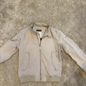 Beige Vår/Höst jacka från J.Lindeberg - Stilig beige jack ifrån J.Lindeberg. Använd 2 gånger och är i exptremt bra skick, storlek XL. Jackan köptes i butik nypris ca 2500kr. Ställ gärna frågor om varan, bilder osv.. Kvitto medföljer tyvär ej.