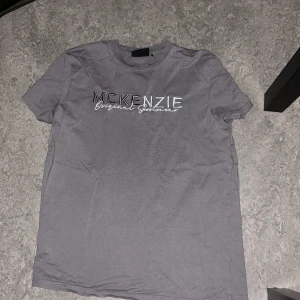 Grå t-shirt från McKenzie i bomull - Snygg grå t-shirt från McKenzie med broderad logga på bröstet i svart och vitt. Klassisk rund hals och korta ärmar. Tillverkad i 100% bomull för en skön och avslappnad känsla. Perfekt för en chill och stilren look.