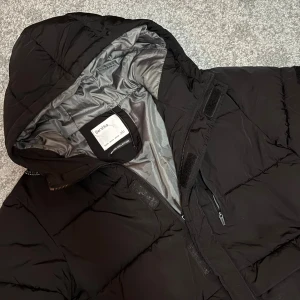 Svart vinterjacka från Bershka - Säljer en svart pufferjacka från Bershka med huva och dragkedja i storlek XS. Jackan har en quiltad look, flera fickor med dragkedja och en snygg, enkel design. Perfekt för kalla dagar och riktigt skön att bära. Fodret är i silvergrått och jackan har en normal passform.