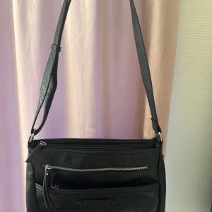 Svart axelväska i skinn med dragkedjor - Stilren svart axelväska i skinn med justerbar rem och flera praktiska fack med dragkedjor. Väskan har en slät, lätt glansig yta och silverfärgade detaljer. Perfekt för dig som vill ha en enkel men snygg väska med plats för det viktigaste.