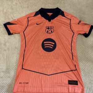 Barça Lamine Yamal Nike fotbollströja - Säljer en FC Barcelona fotbollströja från Nike i orange med mörkblå detaljer. Tröjan har korta ärmar, klubbmärke och sponsortryck på bröstet samt Lamine Yamal och nummer 10 på ryggen. Tillverkad i lätt funktionsmaterial med Dri-FIT-teknologi.