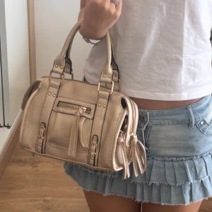 Beige handväska med dragkedja och detaljer - Snygg beige handväska i skinnimitation med både handtag och axelrem. Väskan har coola spänndetaljer, dragkedja framtill och dekorativa tofsar. Perfekt storlek för att få plats med allt du behöver och passar till många olika outfits.