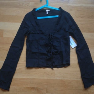 Svart blus med snörning från Nelly - Svart blus från Nelly i 100% bomull med långa ärmar, v-ringning och snygg snörning framtill. Blusen har volangdetaljer längs ringningen och dekorativa sömmar på framsidan. Blusen är genomskinlig.