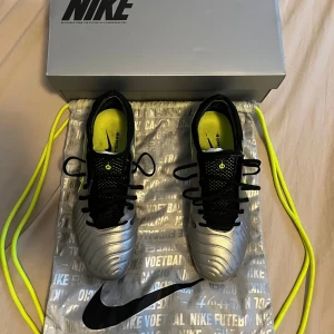 Nike tiempo legend storlek 42 elite  - Säljer ett par Nike fotbollsskor i silver med svarta och neongula detaljer. Skorna har snörning och rund tå, samt dobbar för spel på gräs. Snygg design med Nike-logga på sidan och insida. Perfekt för dig som vill sticka ut på planen!