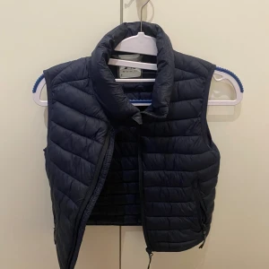 Mörkblå dunväst från McKinley - Snygg mörkblå dunväst från McKinley med quiltad design och hög krage. Västen har dragkedja framtill och två praktiska fickor med dragkedja. Perfekt att slänga på sig när det är kyligt ute. Stilren och enkel att matcha med olika outfits.