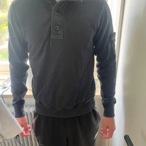 Svart långärmad tröja Stone Island - Svart långärmad tröja från Stone Island med klassisk patch på vänster ärm. Tröjan har en hög krage med tre knappar framtill och ribbade muddar vid ärmslut och nederkant. Materialet är mjukt och känns som bomull, perfekt för en chill och stilren look.