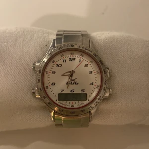 Silverfärgad klocka från Gul - Stilren klocka från Gul med rund urtavla i vitt och digital display. Klockan har en silverfärgad länkarmband i metall och flera knappar på sidan för extra funktioner. Urtavlan har tydliga siffror och en röd sekundvisare. Perfekt för dig som gillar sportig design.