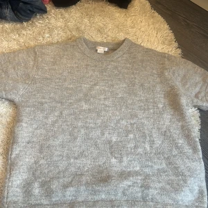 Grå stickad tröja från Gina Tricot - Mysig grå stickad T-shirt från Gina Tricot med rund hals och ribbade muddar. Perfekt för kyliga dagar och enkel att matcha med jeans eller kjol. Tröjan har en klassisk, normal passform och är tillverkad i mjukt material som känns skönt mot huden.